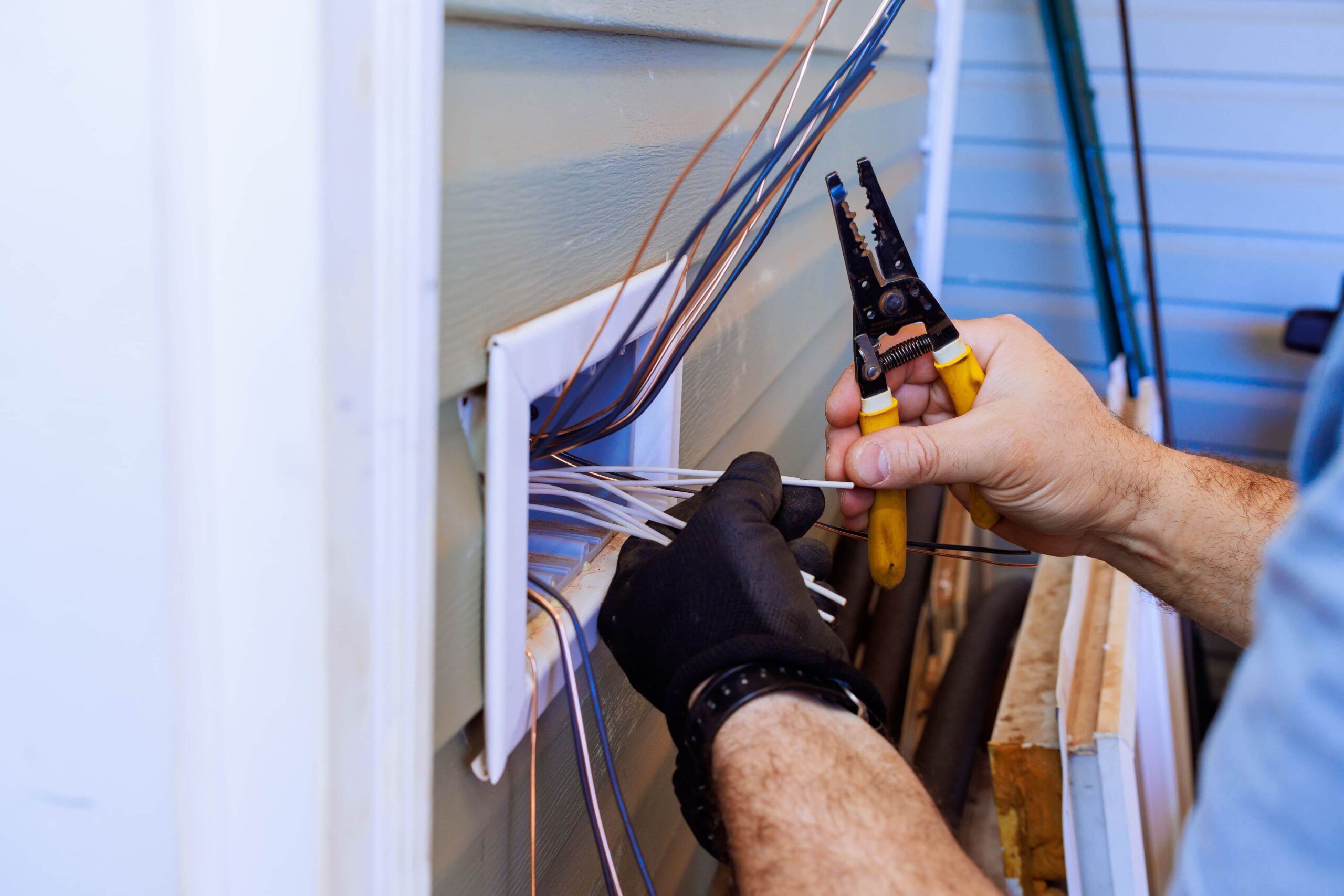 Close-up of a technician's hands in black protective gloves using yellow-handled wire strippers to prepare electrical cables at an exterior wall outlet box.