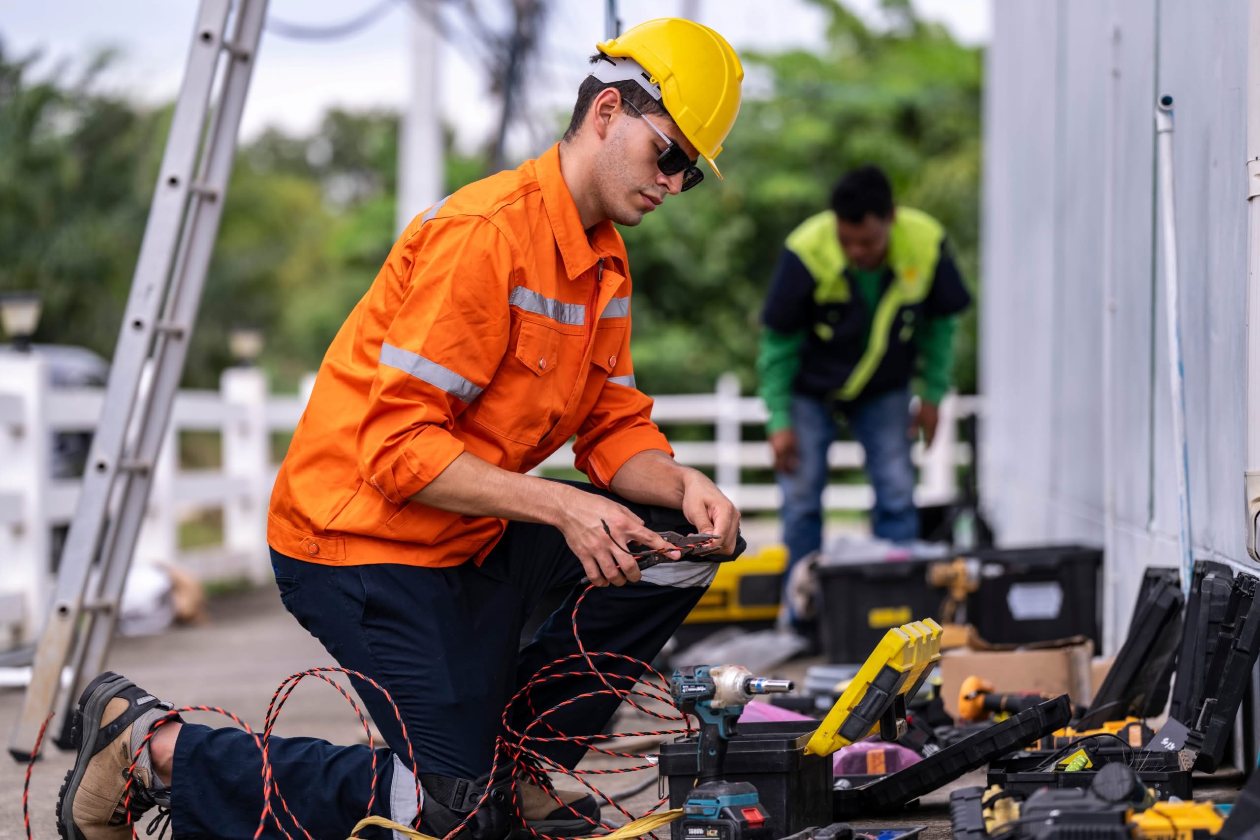 Commercial electrical maintenance in Eustis Florida 1 A technician kneeling on the ground wearing a bright orange high-visibility safety jacket, yellow hard hat, and sunglasses, carefully stripping or splicing wires with hand tools surrounded by open toolboxes and power equipment.